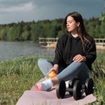 Woman sitting on yoga mat beside a lake with exercise equipment, enjoying a calm outdoor workout.